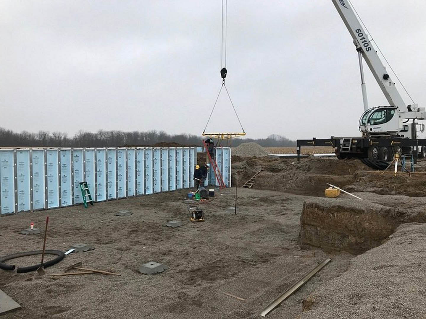 Crane hoisting metal beam at lakeside construction site, ladder leaning against unfinished wall, flatbed truck with white crane, cloudy sky overhead