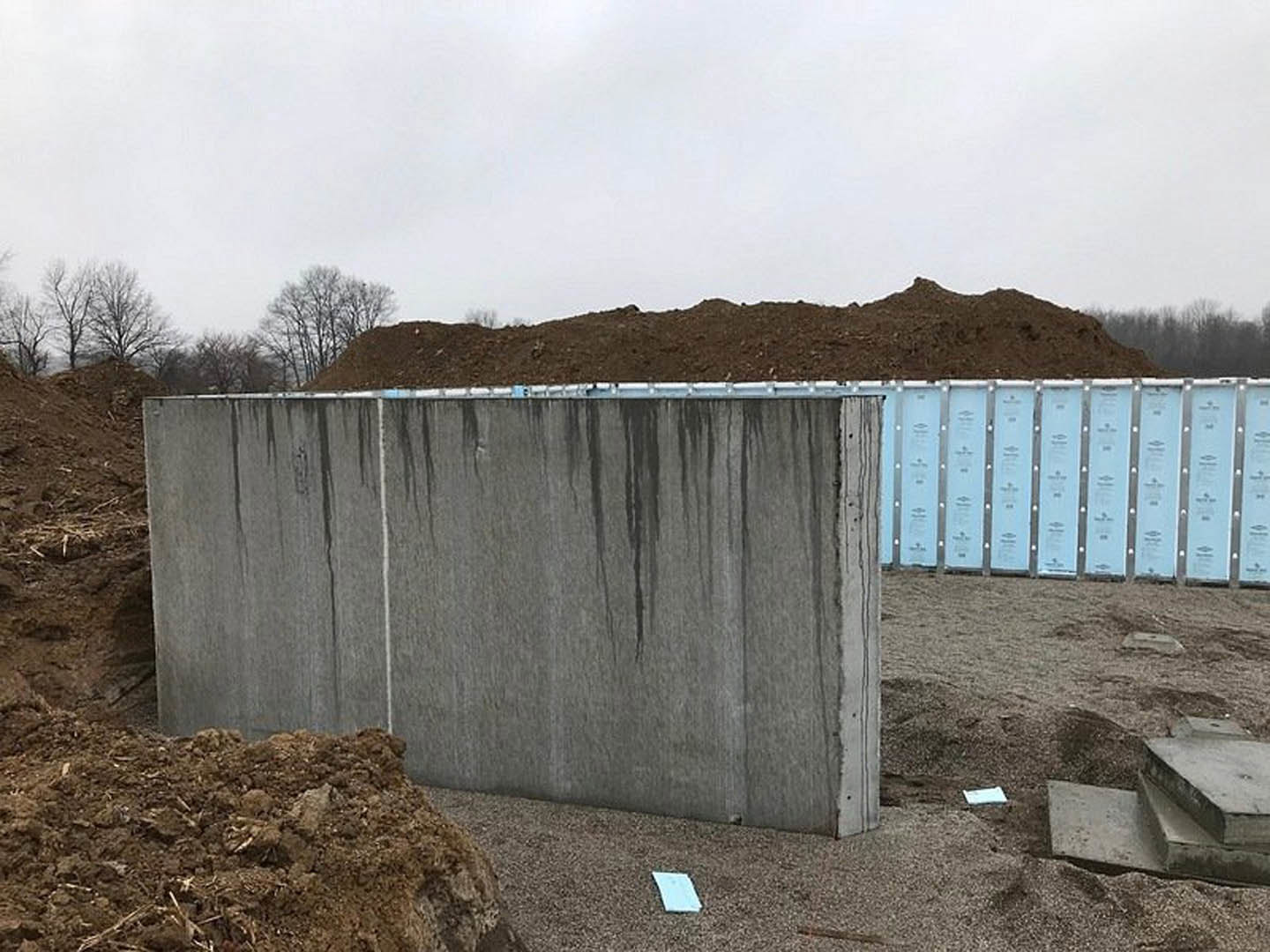 Smooth concrete wall at a residential construction site, bordered by a mound of dirt and straw, under a cloudy sky.