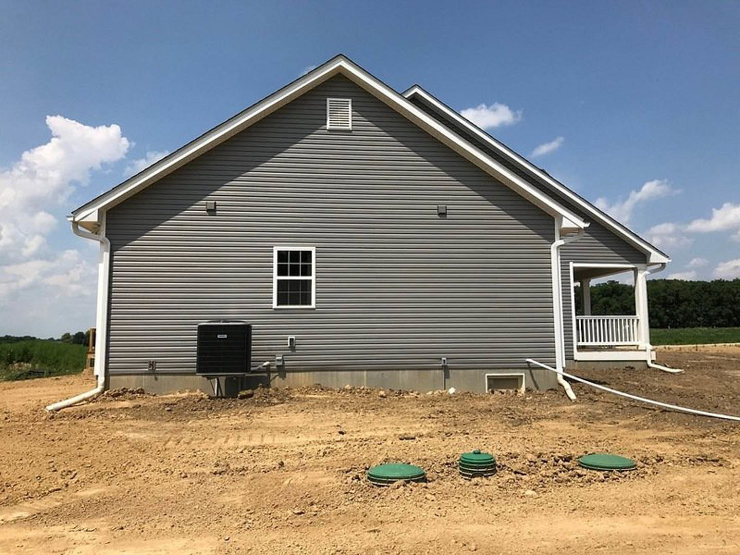 Partially built house with white siding, multi-pane window, white wall vent, black square utility box, green container on dirt lot, blue sky overhead