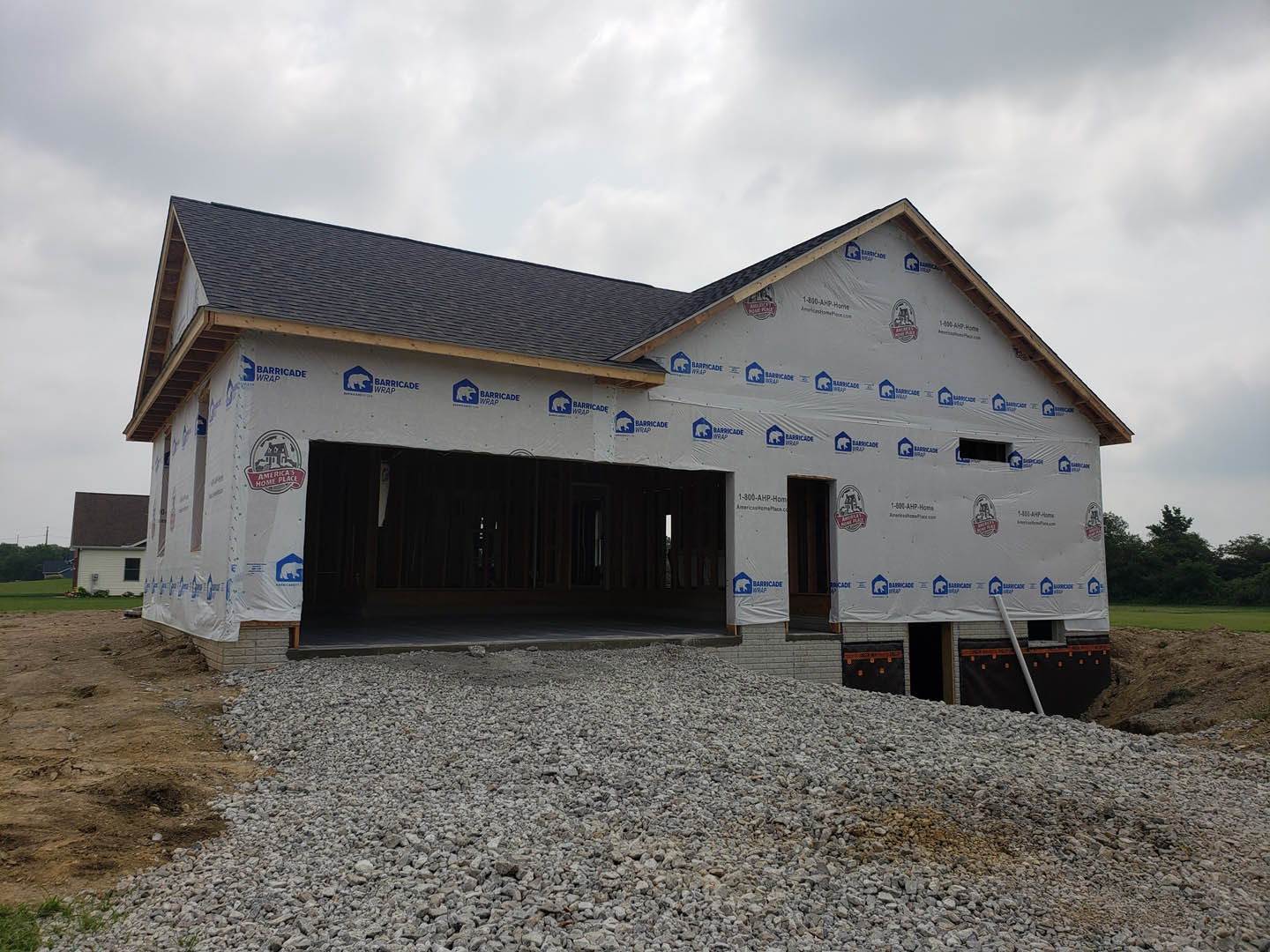 Two-story house under construction with white plastic sheeting covering exterior walls, attached garage, brick retaining wall, pile of rocks in front, cloudy sky overhead
