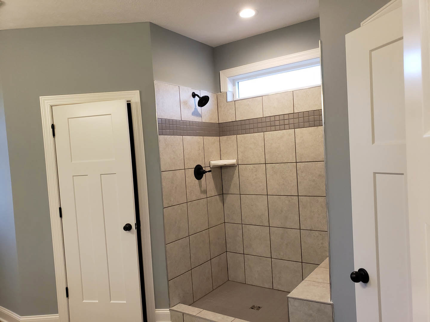 Bathroom with tiled shower, glass shower door, white paneled door with black knob, ceiling light, window above shower, and wall-mounted mirror.