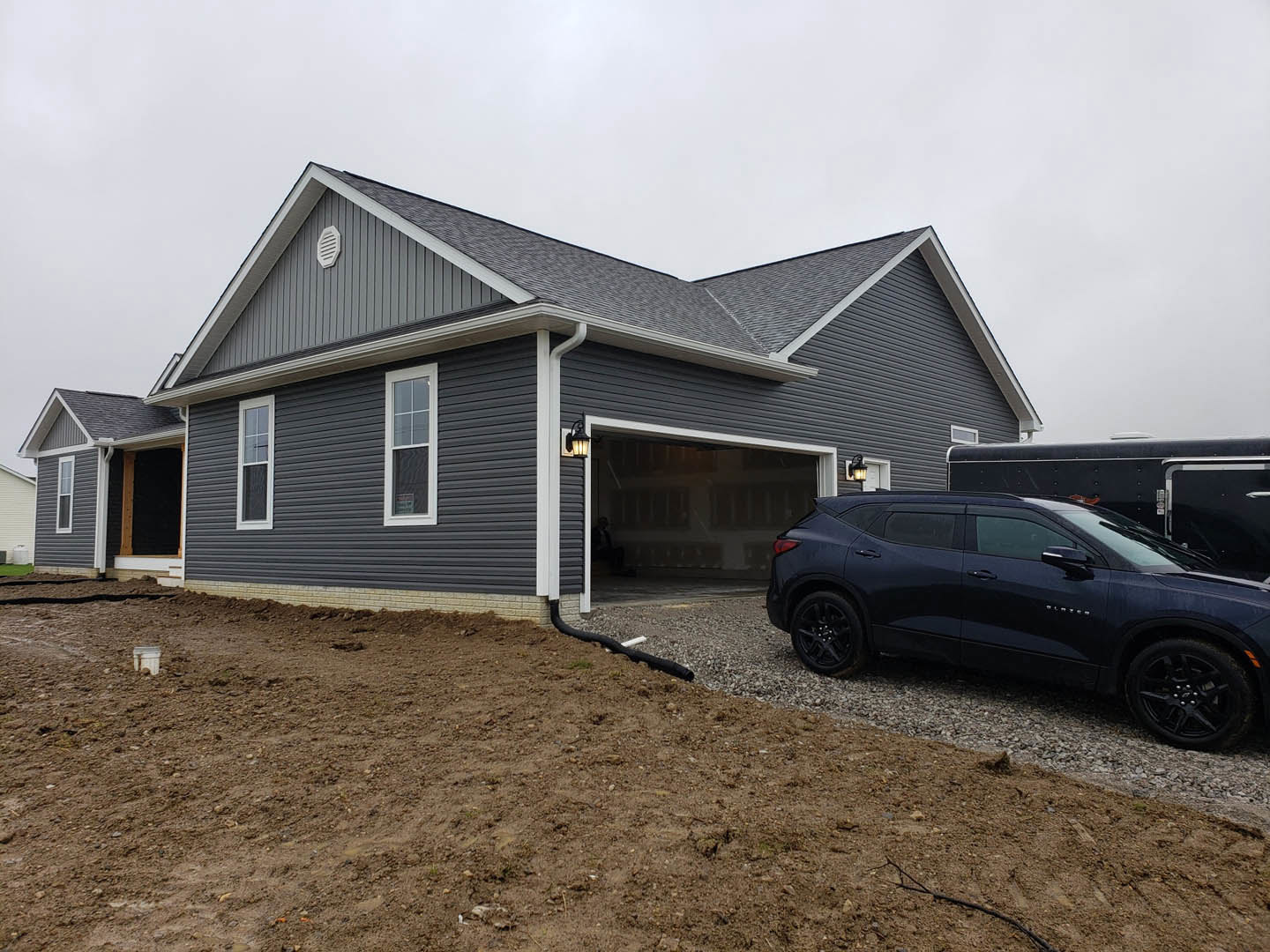 Gray sedan parked on gravel driveway beside white-framed window and attached garage, dirt hill with black drainage pipe visible near house, enclosed trailer parked adjacent to car