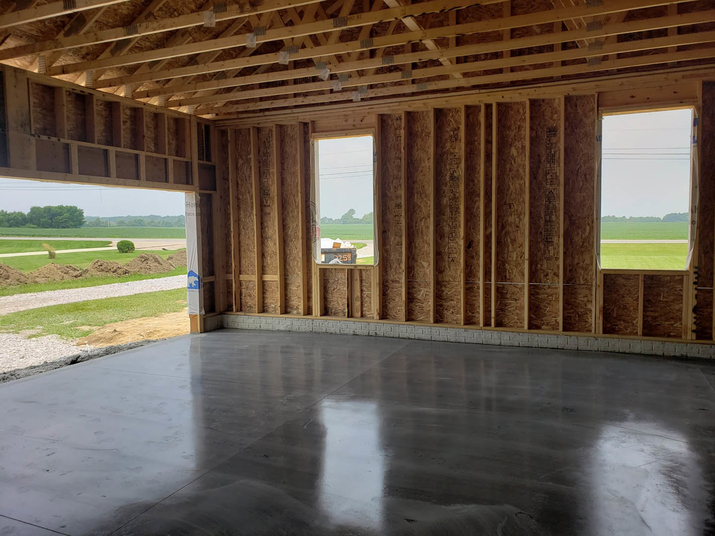 Concrete floor with reflective grey finish, exposed brick wall, wooden roof trusses with construction tape, large windows overlooking grassy field and distant trees