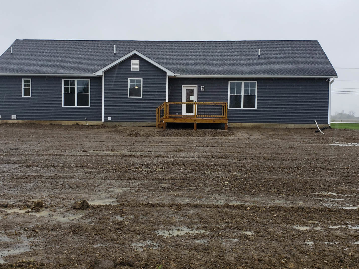 Two-story house with light siding, wooden deck and railing, white-framed windows, dirt yard in foreground, open sky above