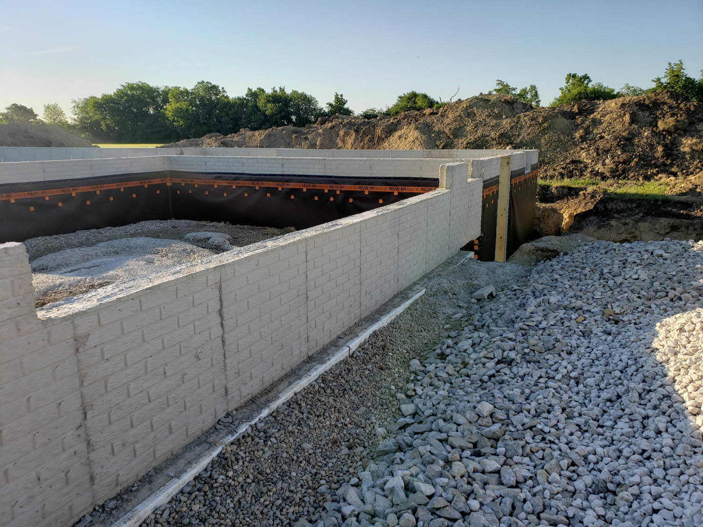 Brick wall foundation with gravel base, black and orange construction tape, pile of rocks beside the wall, clear blue sky and trees in background