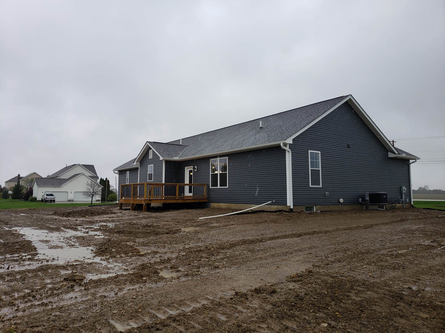 Wooden deck with railing attached to a cottage-style house, rain gutter along roof edge, large window, tire tracks on bare dirt yard, overcast sky, sparse grass and trees in