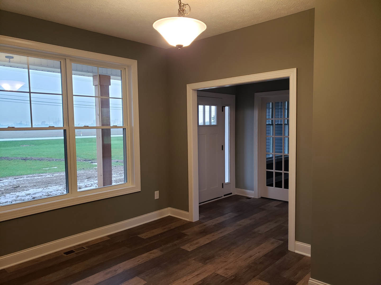 Wood flooring with white trim, large window overlooking grassy field, white door with black handle, ceiling light fixture, neutral walls