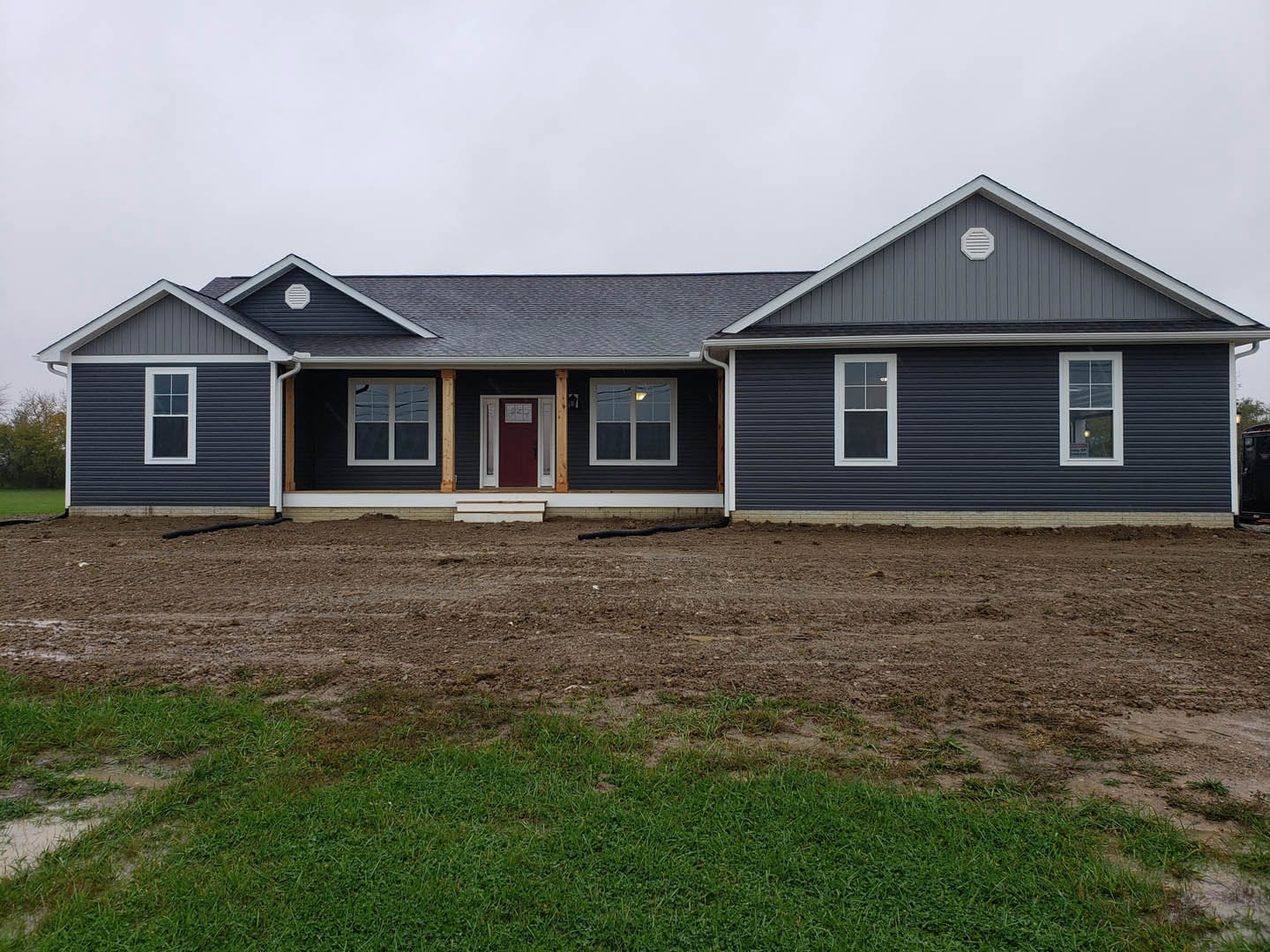 Two-story house with gray siding, red front door with white trim, white-framed windows, dirt yard in foreground, garden hose visible in background