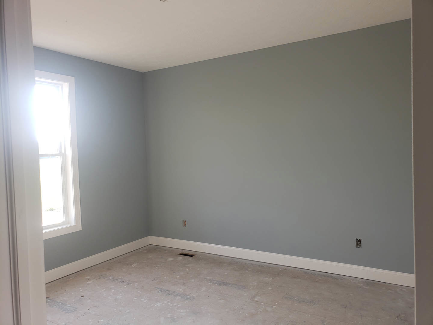 Sunlit room with white plaster walls, large window, grey ceiling, and unfinished flooring with visible holes