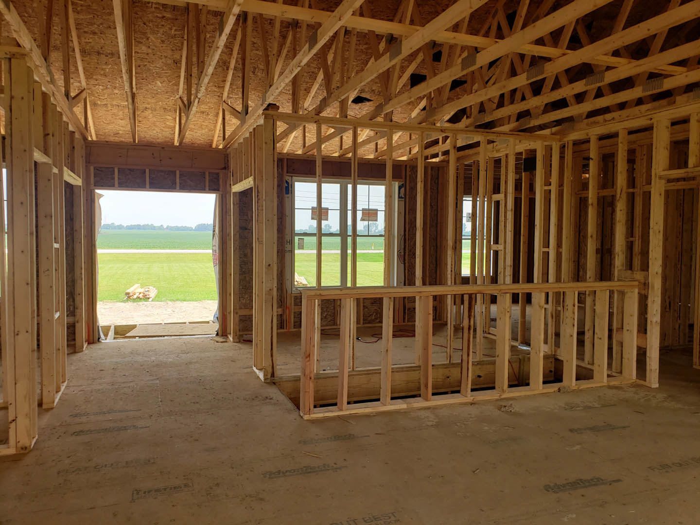 Wood-framed room under construction with exposed ceiling beams, large window overlooking grassy field, unfinished floor with lumber planks.