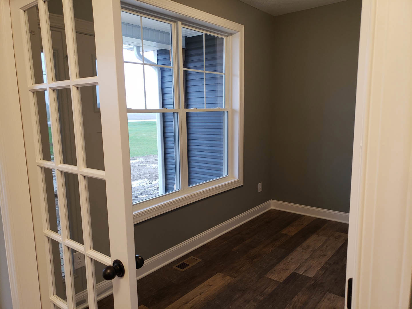 White-trimmed window above vent on natural wood floor, black door knob on white wall, open door leading into bright room