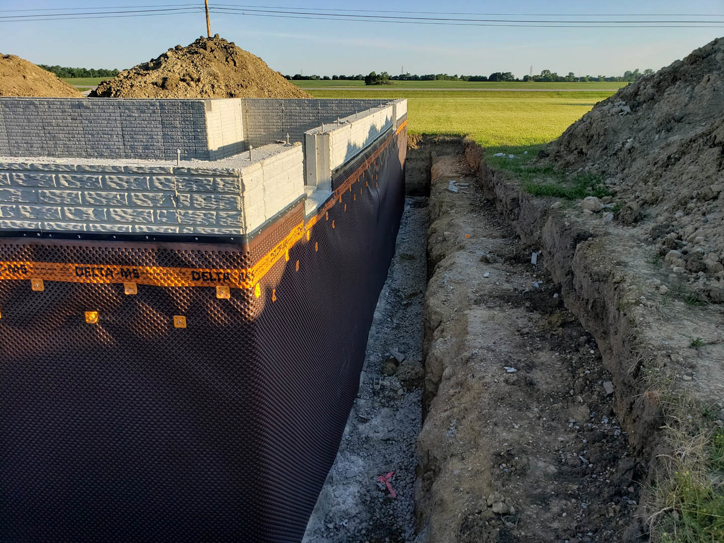 Concrete foundation with exposed rebar, dirt piles, and construction materials; brick and stone wall sections visible; power lines overhead; pink ribbon marking area on grey