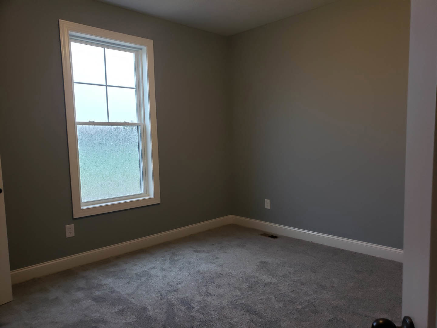 Grey carpeted room with white plaster walls, large window featuring frosted glass, and overcast sky visible outside