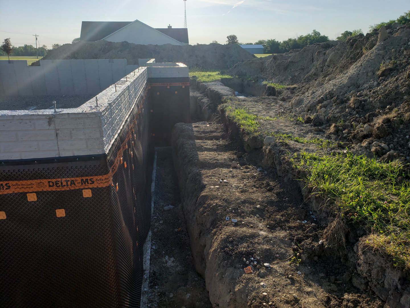 Wide trench dug in brown dirt field with brick wall lined in orange and black tape, white house with dark roof in background, construction sign and metal tower visible, cloudy sky