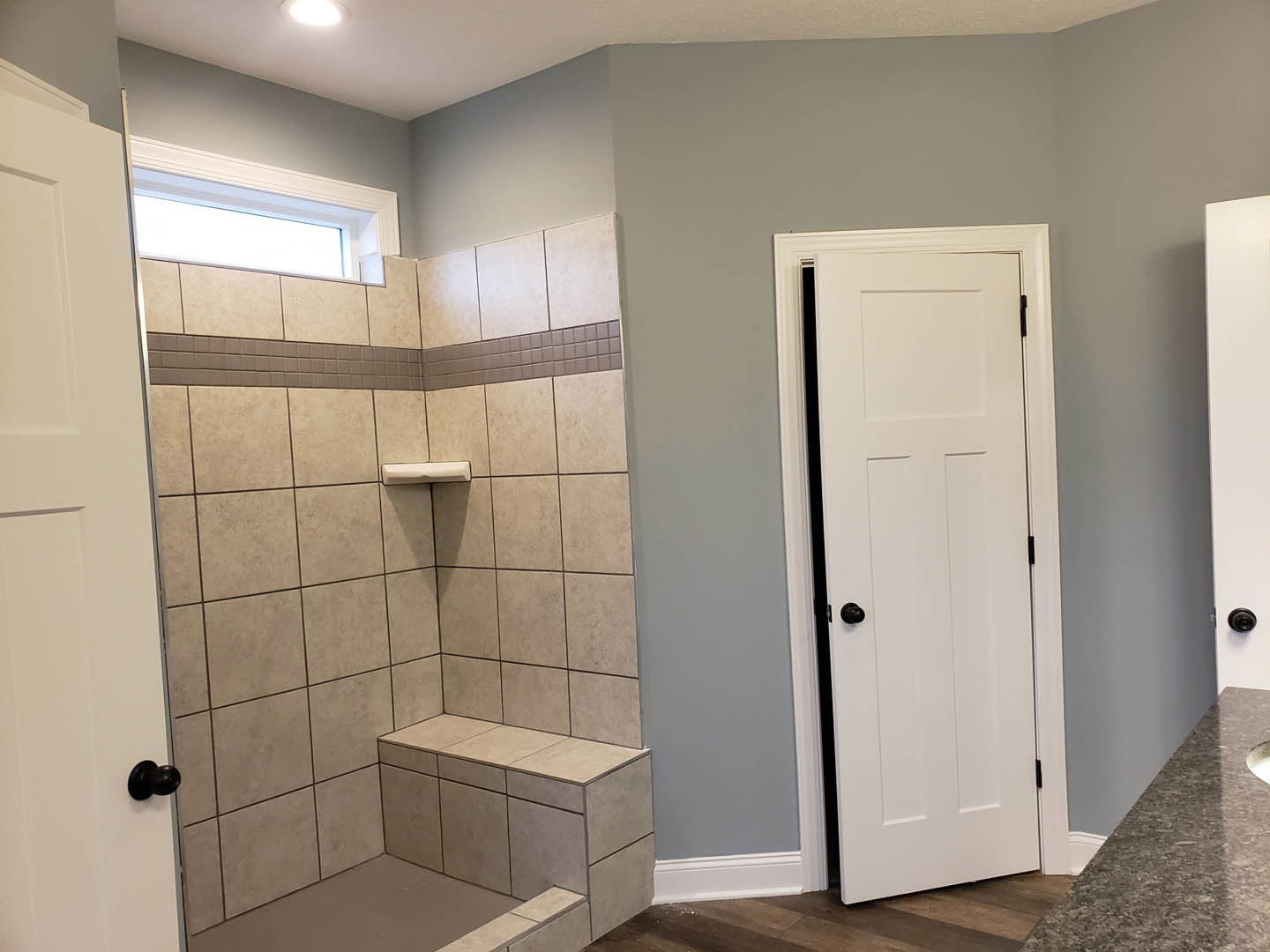 Bathroom with marble countertop, tiled shower step, white cabinetry, and white door featuring a black knob
