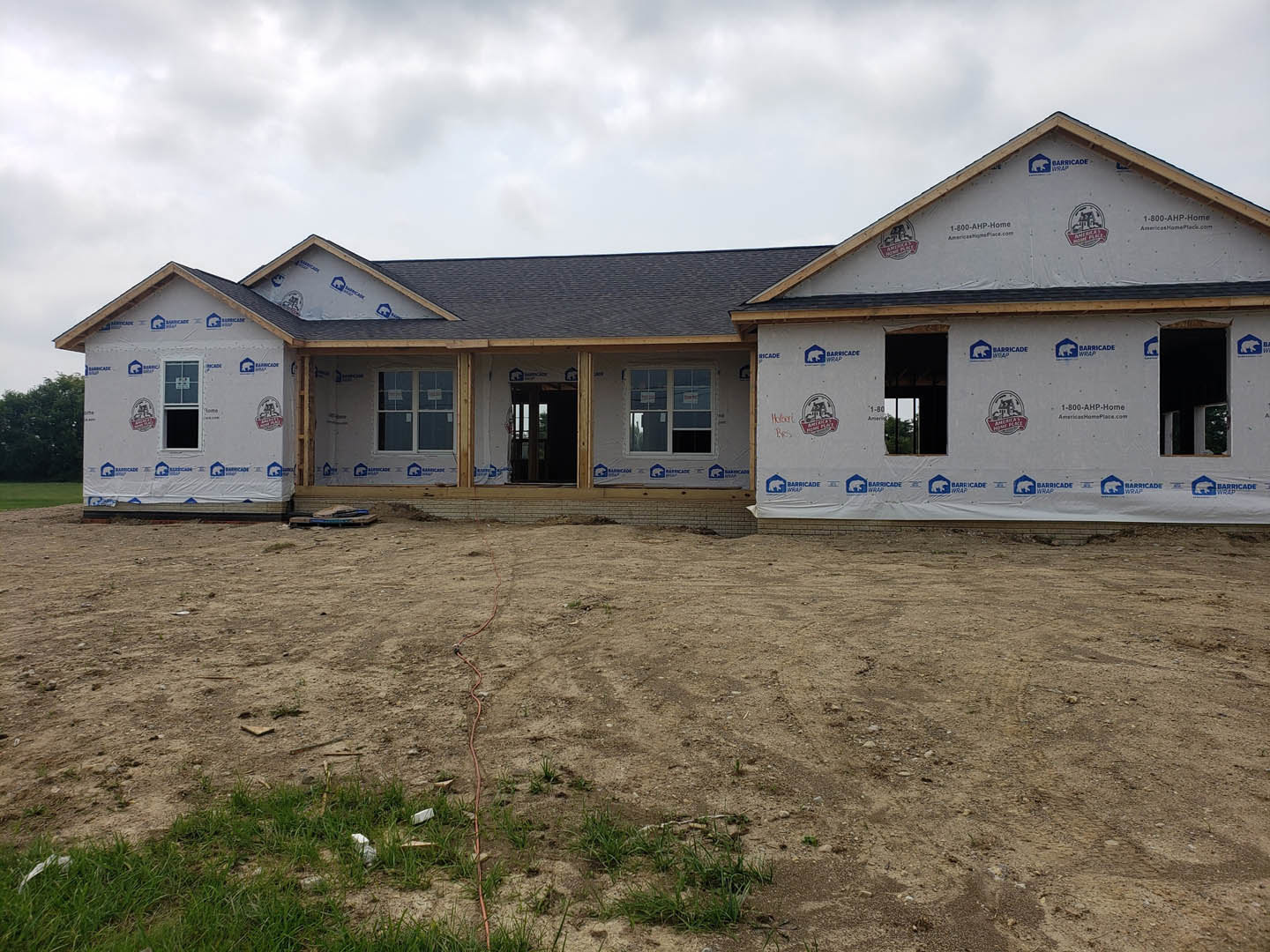 Two-story house under construction with exposed framing, white exterior walls, and large windows, set on a dirt field with scattered grass and construction materials.