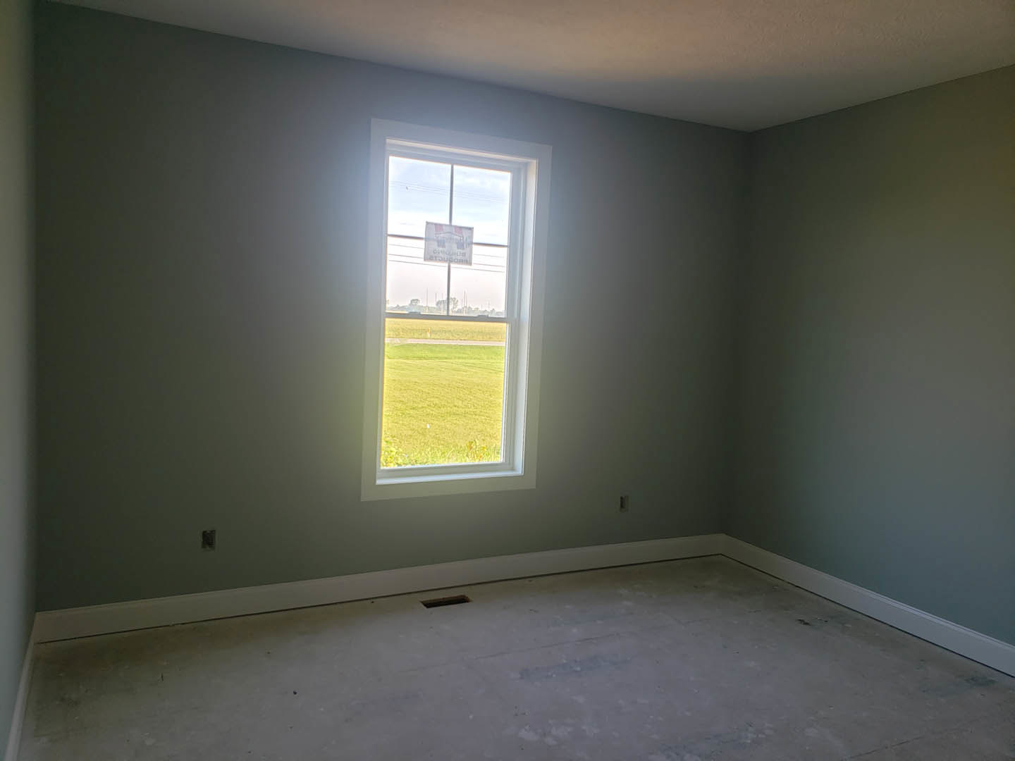 Bright room with white plaster walls, large window overlooking green grass field, light-colored flooring with visible hole, blue accent background, and sign with text displayed on