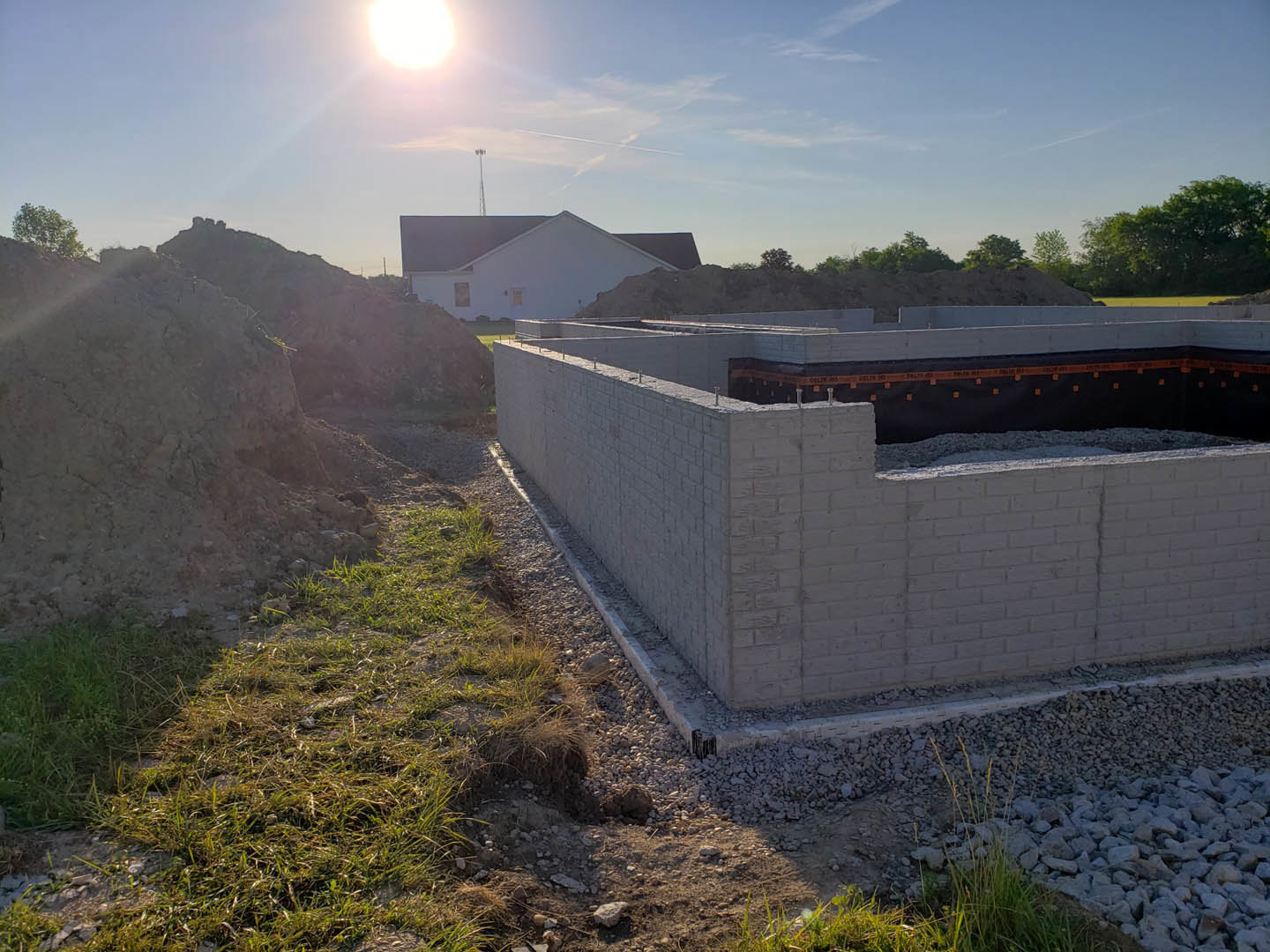 Brick home under construction with exposed walls, black tarp, and roof partially finished; pile of dirt and grassy ground in foreground, blue sky with scattered clouds overhead.