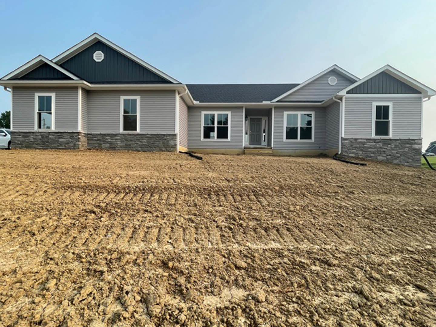 Two-story house under construction with white-framed windows, gray door, and light siding, surrounded by dirt and a small hill in the foreground
