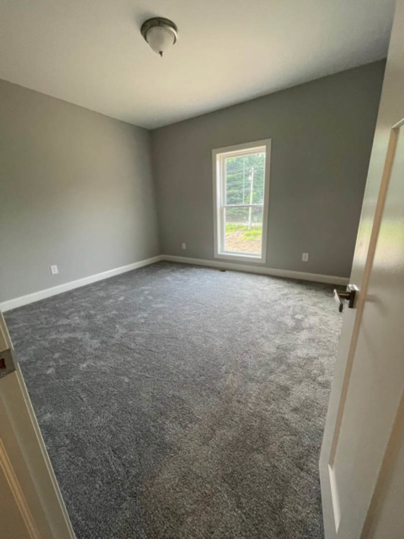 Bedroom with grey carpet flooring, white framed window, white plaster walls, and a close-up of a modern light fixture; partial view of a door with a silver handle.