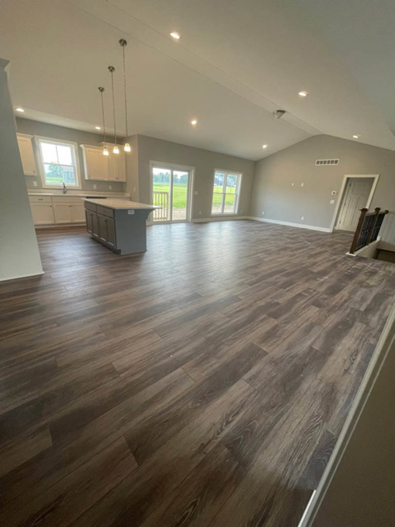 Spacious open floor plan featuring hardwood flooring, white kitchen island with drawers, glass door overlooking grassy yard, and a white door near a staircase
