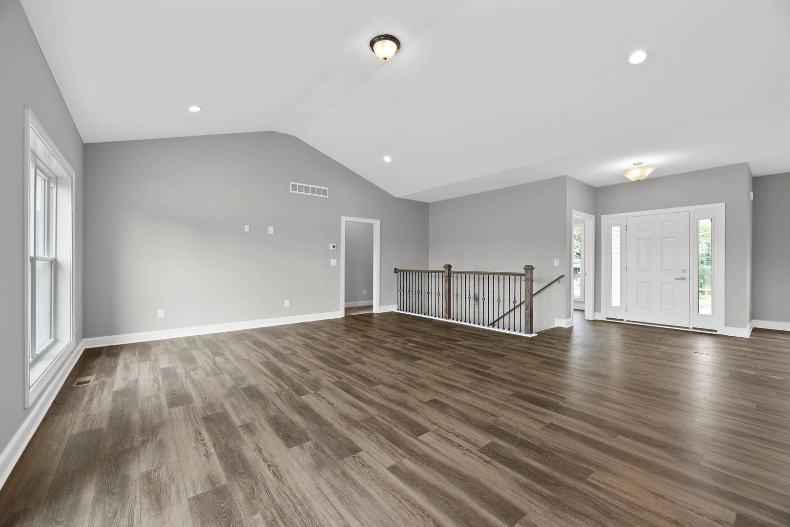 Spacious room with light wood flooring, white door featuring glass panes, metal railing, ceiling light fixture, and neutral grey walls