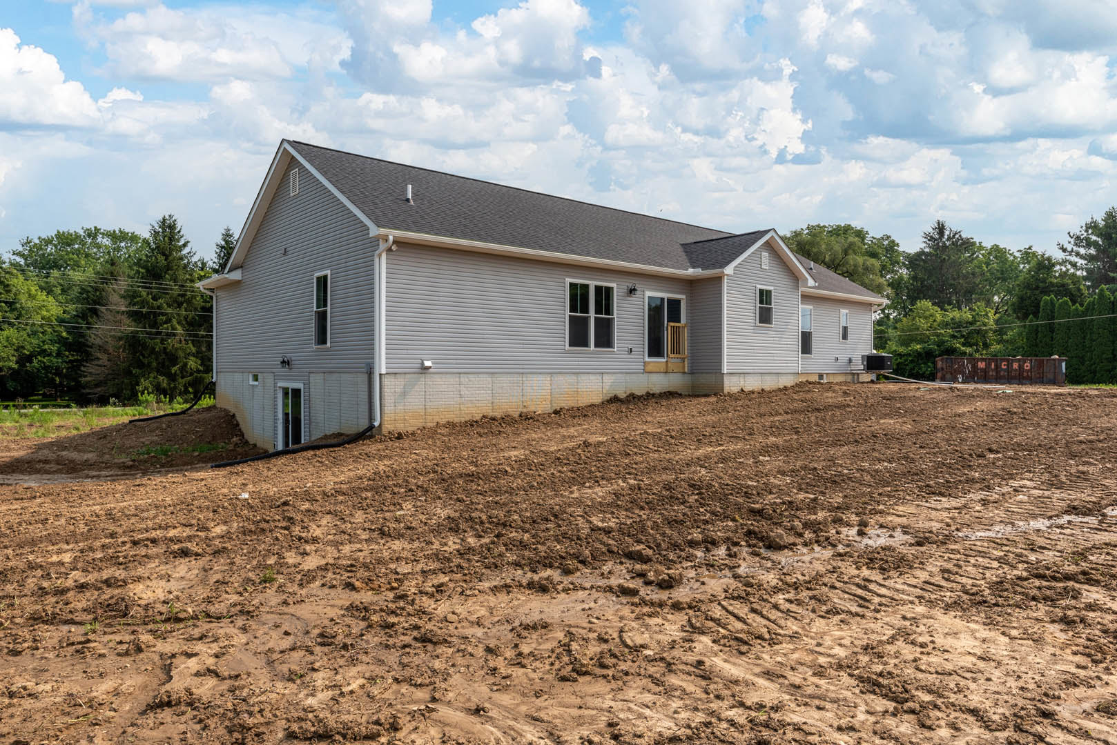 Two-story house with white-framed windows, gray roof, and fenced yard bordered by a dirt hill, muddy ground, and scattered trees under a cloudy sky