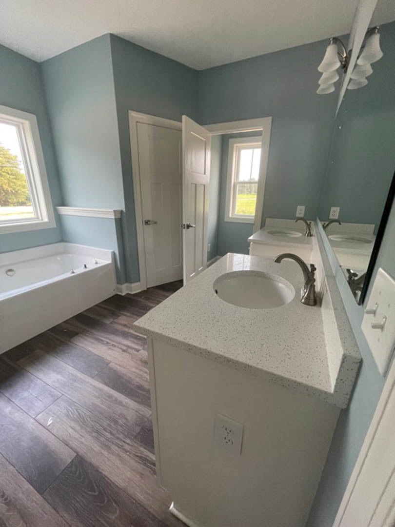 Modern bathroom featuring a freestanding white bathtub with silver fixtures, rectangular sink set in a stone countertop, large window framing leafy tree views, light gray tile