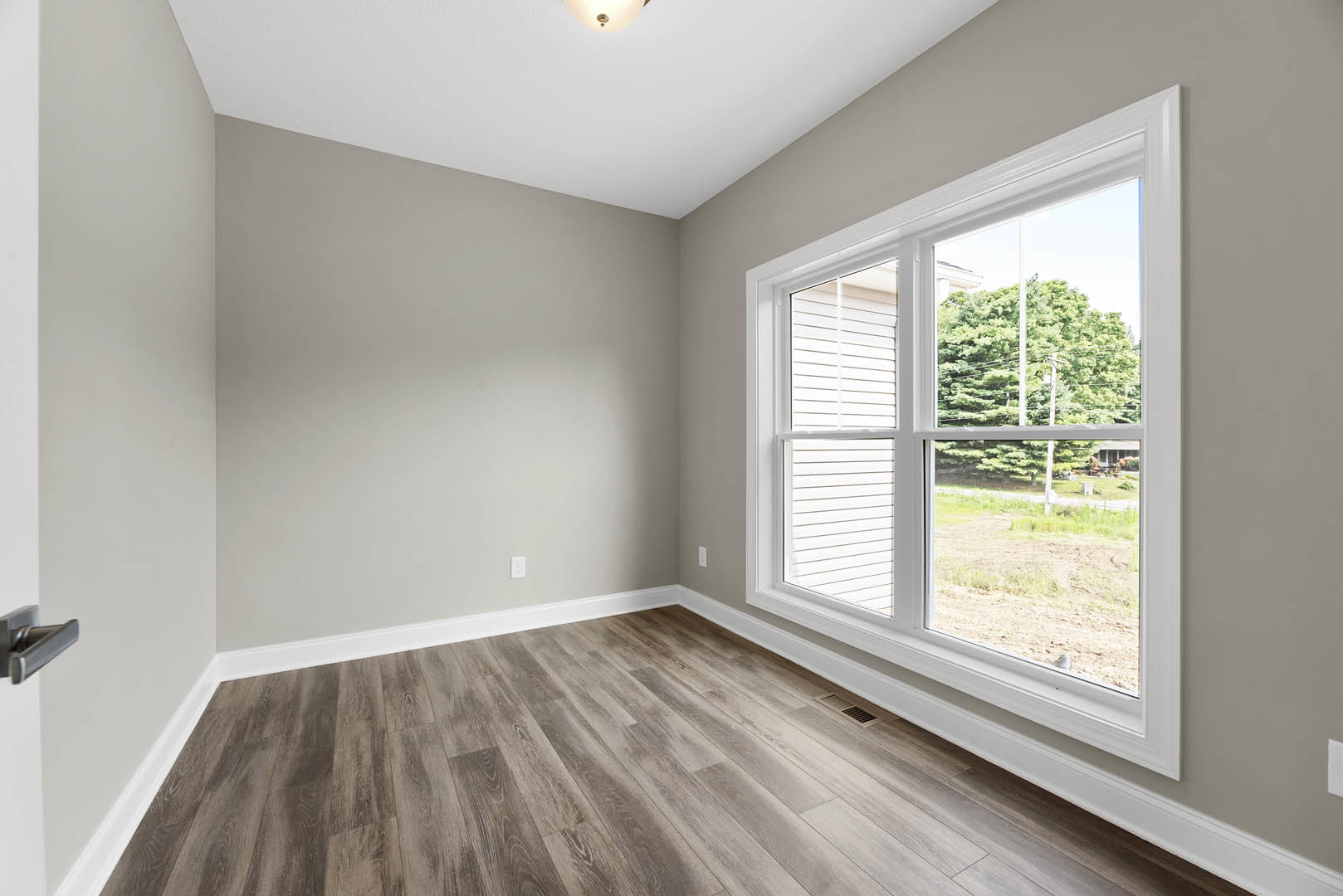 Sunlit room featuring a large window, white plaster walls, and polished hardwood flooring with a close-up of a modern door handle