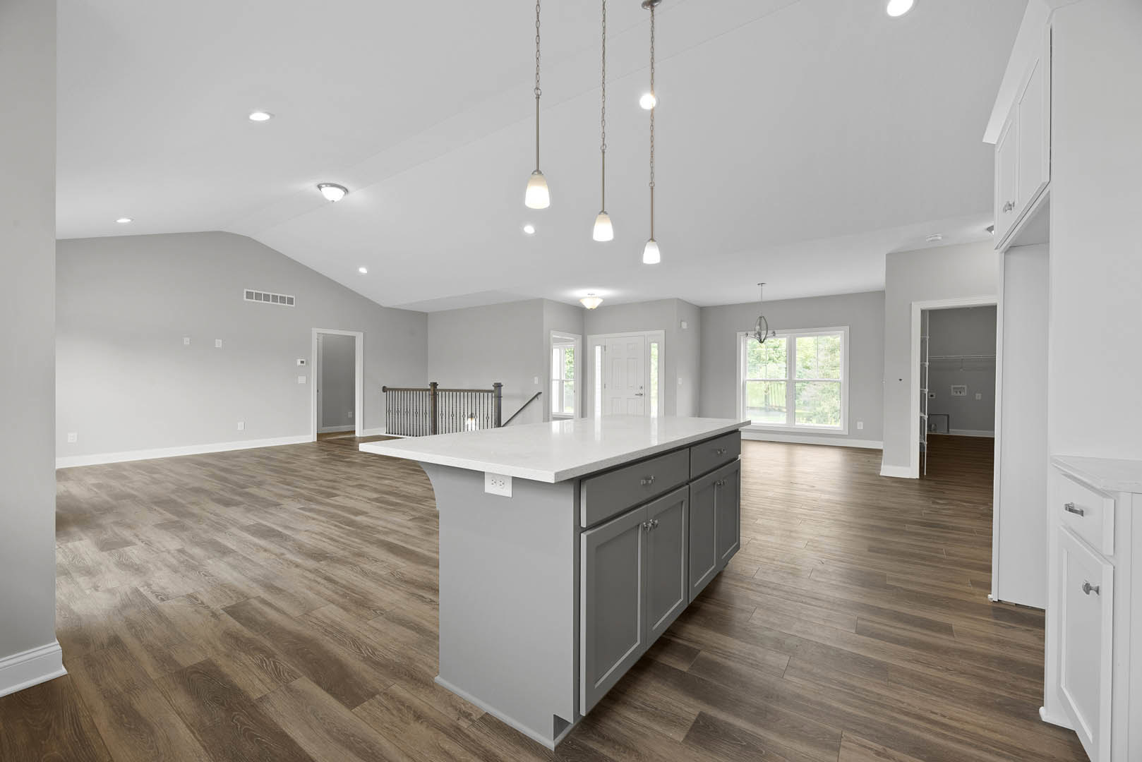 Spacious kitchen featuring a large white island with quartz countertops, hardwood flooring, white cabinetry with silver handles, pendant lighting, and windows overlooking trees.