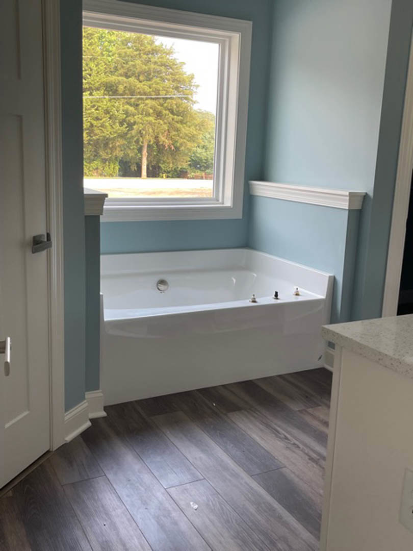 Freestanding white bathtub beside a window with trees visible outside, wood floor with white trim, minimal objects on tub edge, white wall and chrome tap fixtures