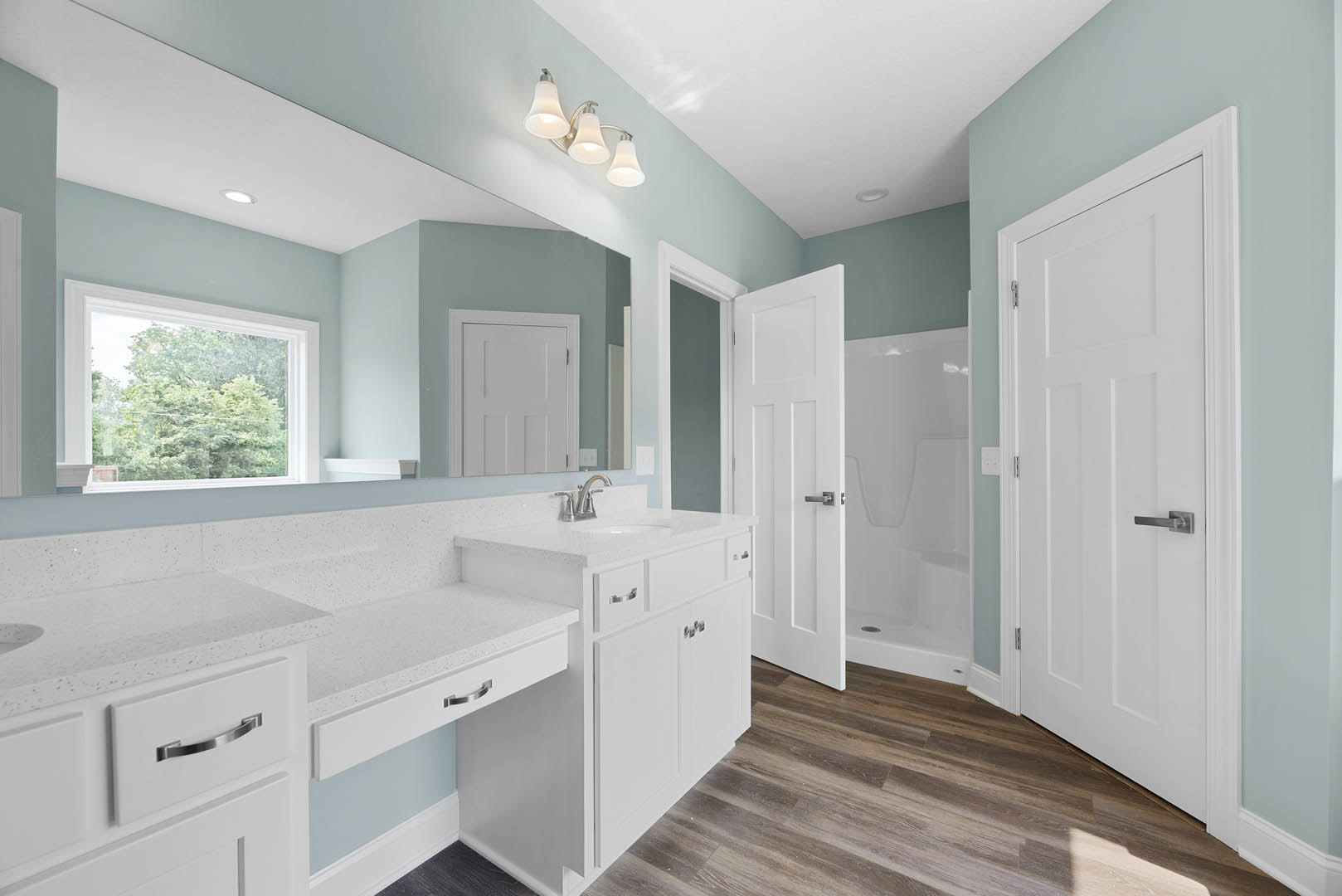 Bathroom with white vanity and undermount sink, large framed mirror above, three-bulb metal light fixture, window with view of green trees, white door with matching trim, neutral