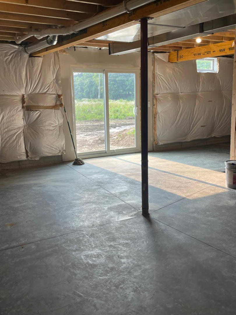 Concrete floor with a metal support pole, white plastic-covered wall, door, and window overlooking a grassy field