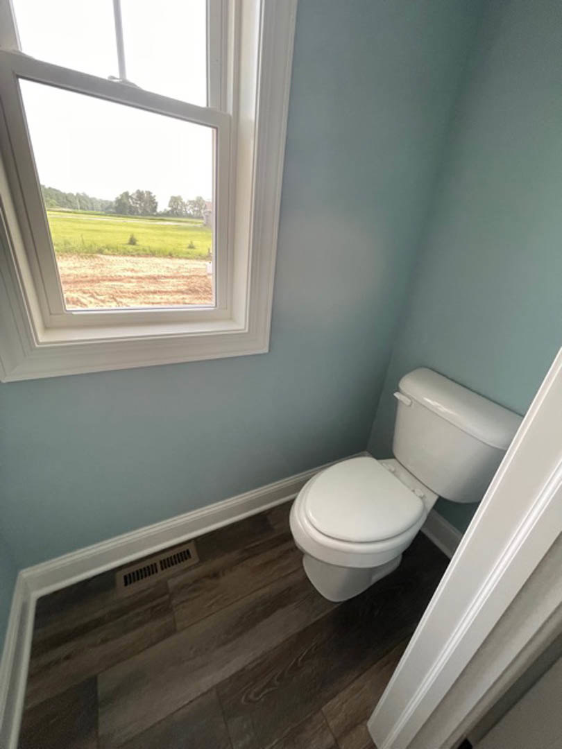 White toilet with lid up and tank, wood floor, wall vent, large window overlooking grassy field, plaster walls