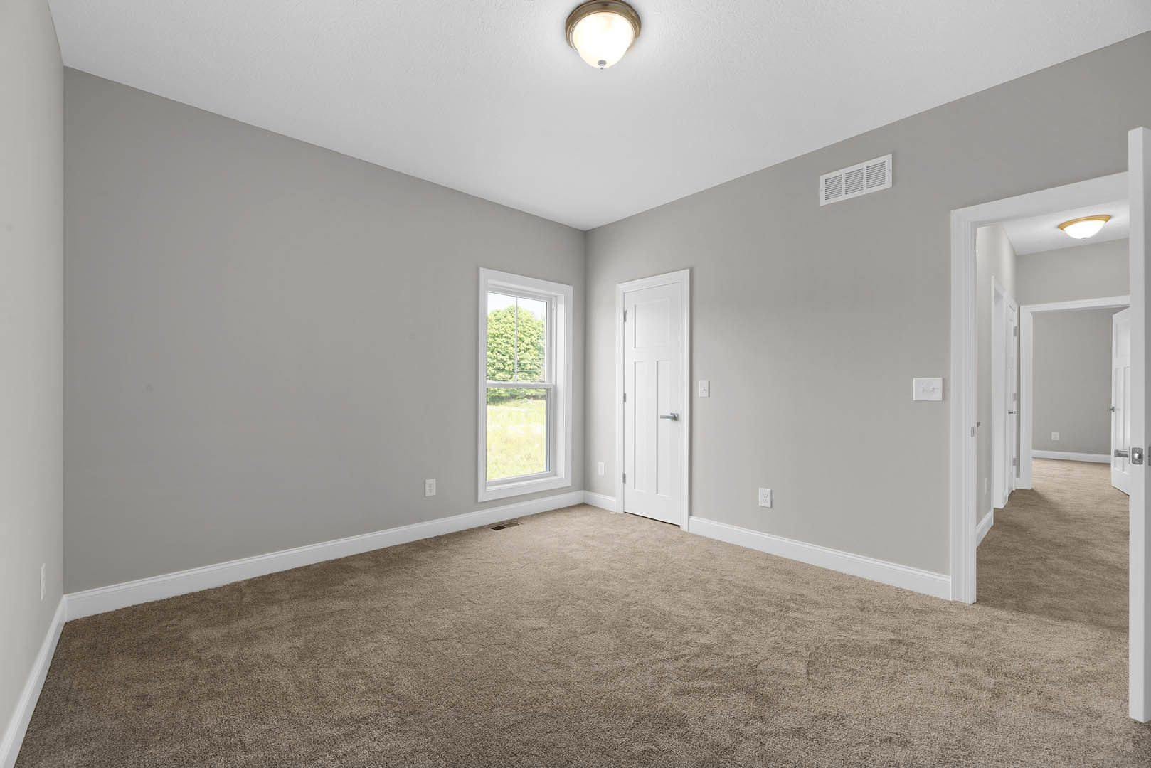 Carpeted room with white door featuring silver handle, large window overlooking green field, ceiling-mounted light fixture, wall vent, and white plaster walls with molding.