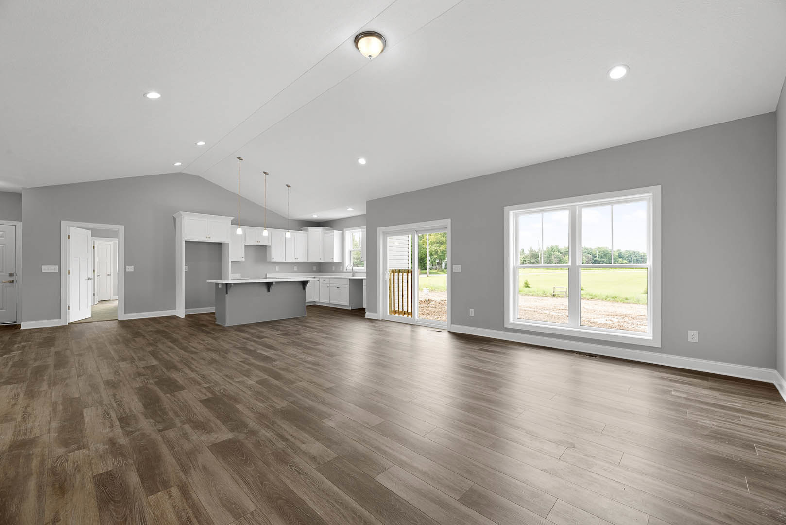 Spacious room with hardwood flooring, open kitchen area, large window overlooking a field, white plaster walls, and ceiling-mounted light fixture