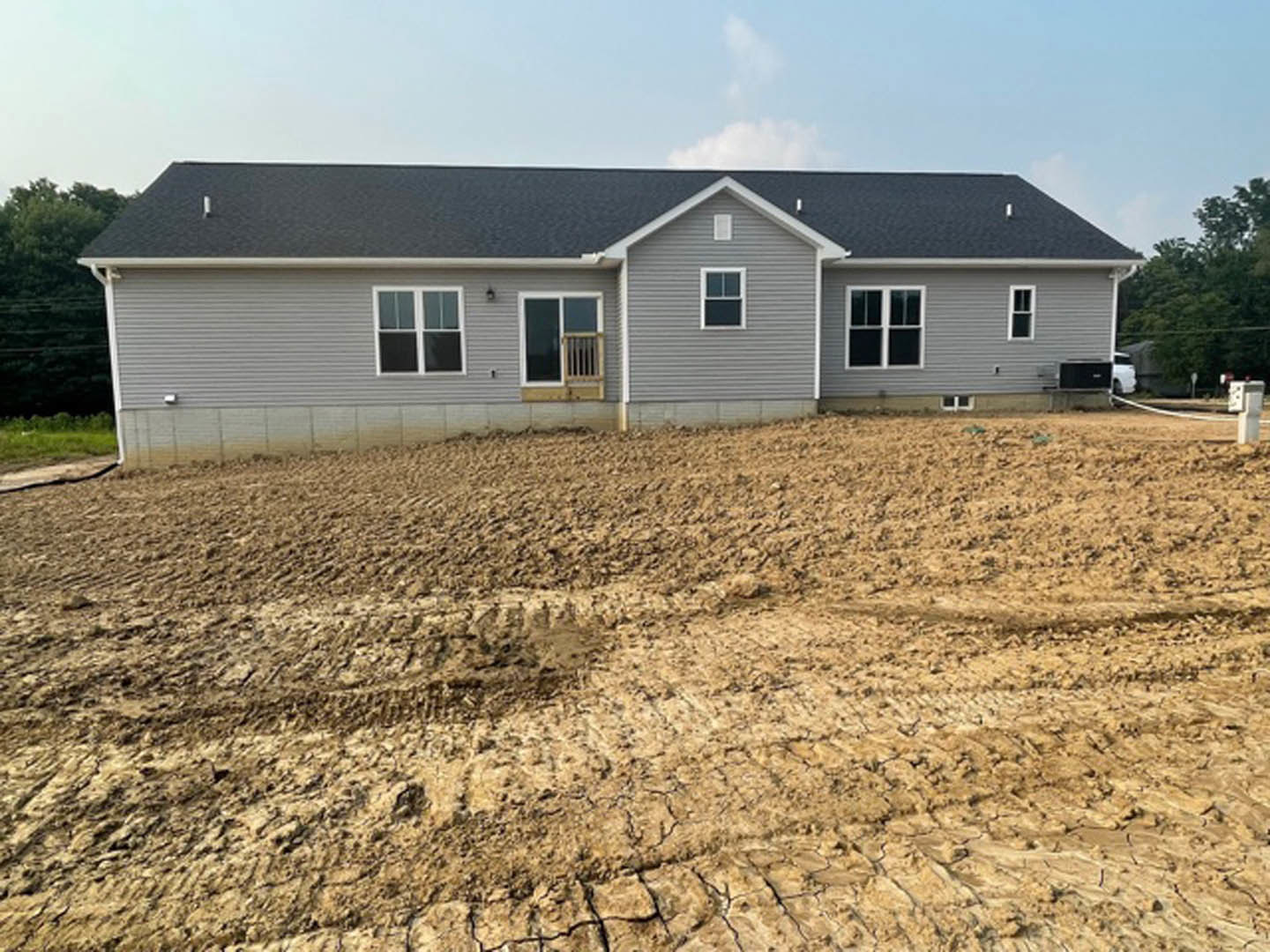 Grey house with white roof under construction, dirt lot with tire tracks, visible windows and door, trees in background