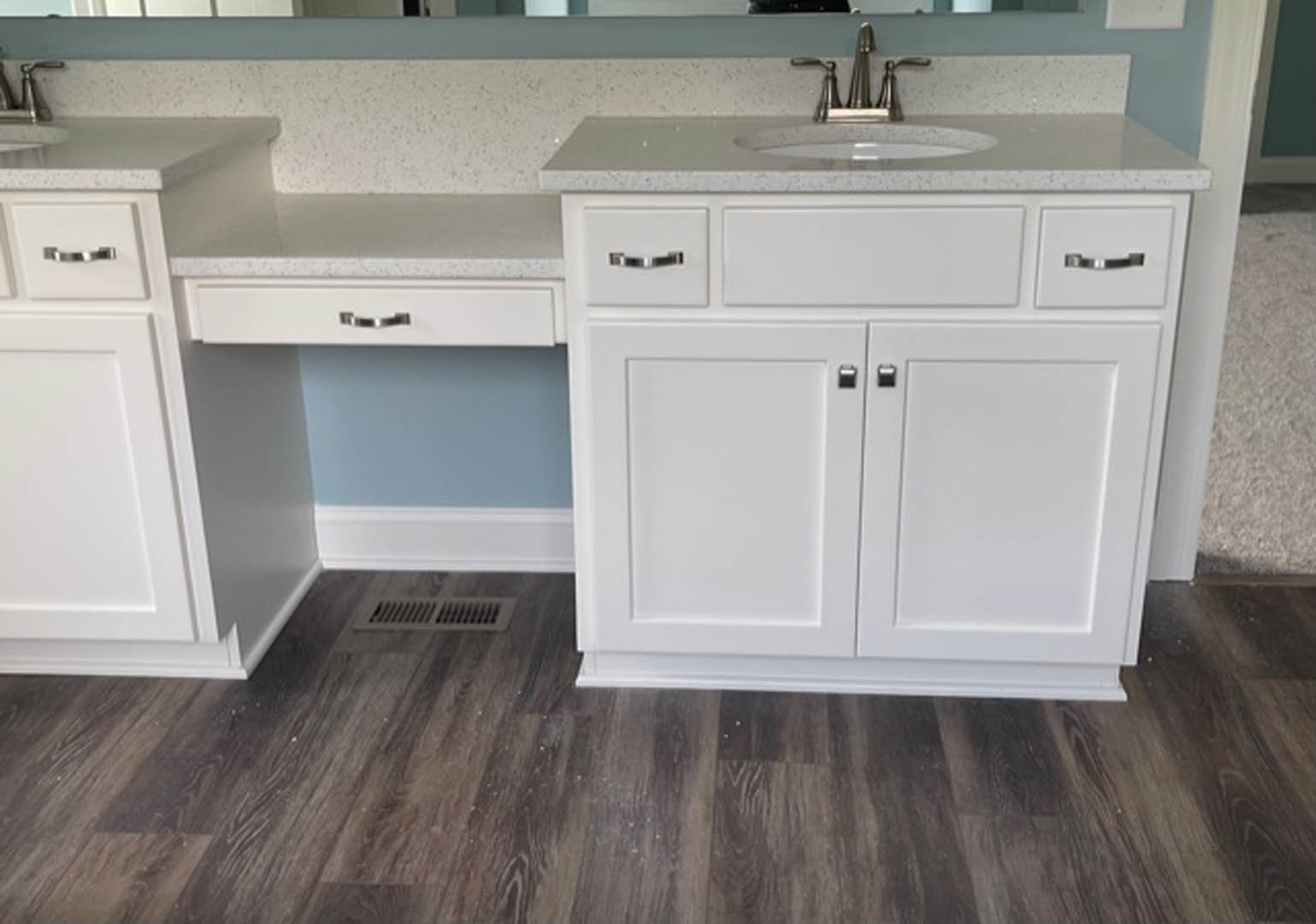White bathroom sink with silver faucet and handles set in a light countertop, wood flooring with white trim, nearby cabinetry and drawers, vent visible on surface, close-up of