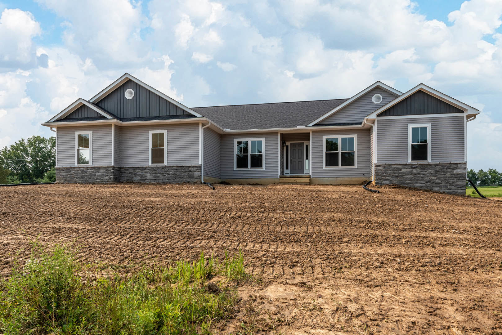 Two-story house under construction with white-framed windows, surrounded by a dirt field with tire tracks, under a clear blue sky