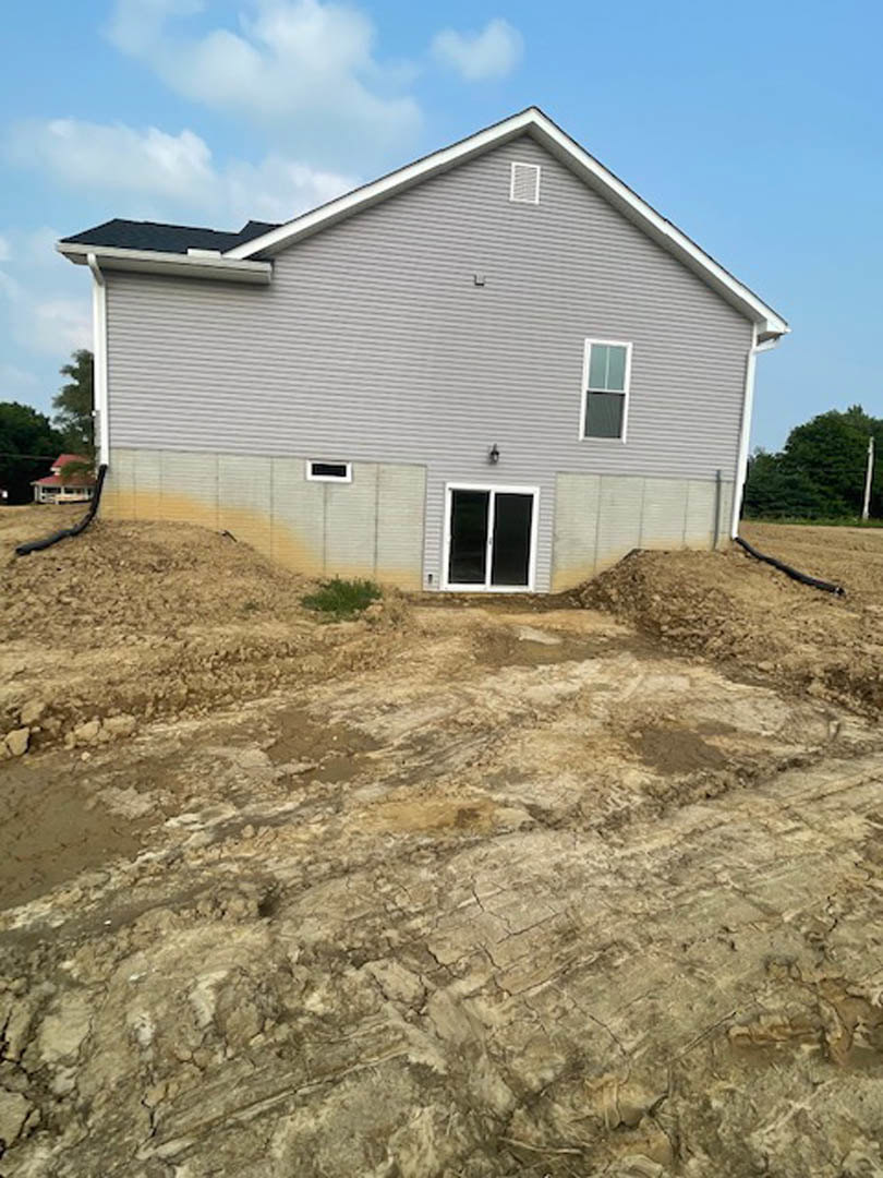 Partially built house with exposed framing, white window frames, dirt piles in front yard, sparse green bush, and clear blue sky overhead