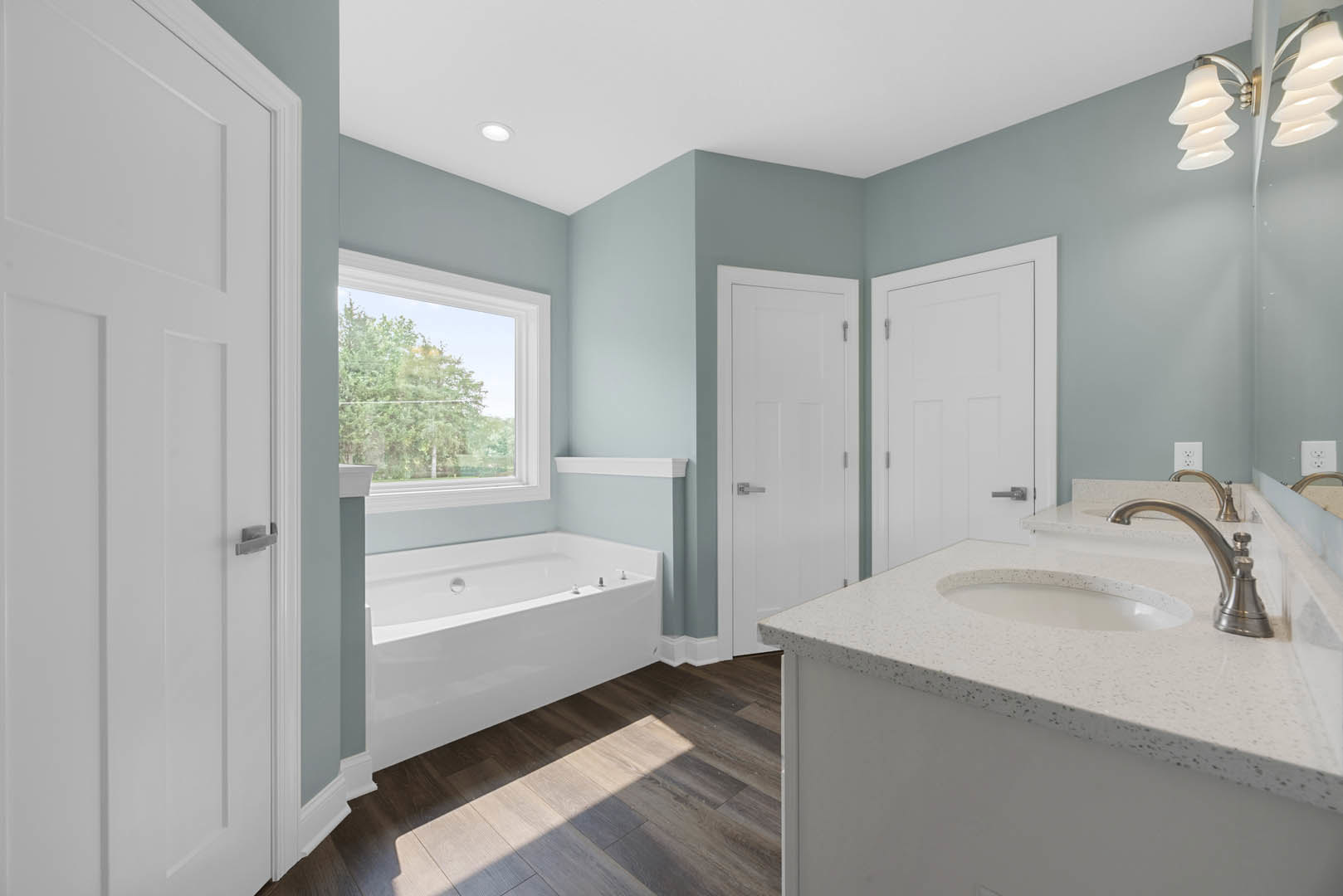 White freestanding bathtub beside a large window with trees visible outside, modern sink and chrome faucet on a vanity, light fixture mounted above, neutral tile walls and floor