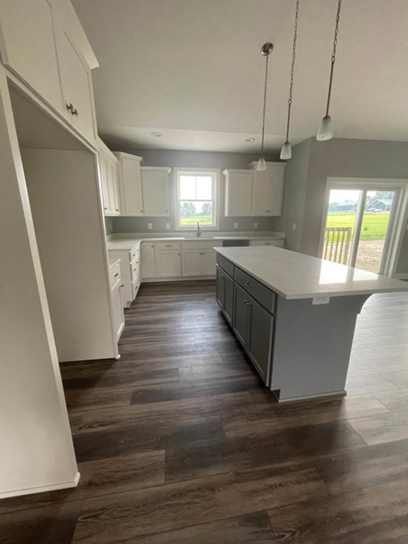 Spacious kitchen featuring a large island with a white countertop, wood flooring, white cabinetry, and a window overlooking trees, grass, and a fence.