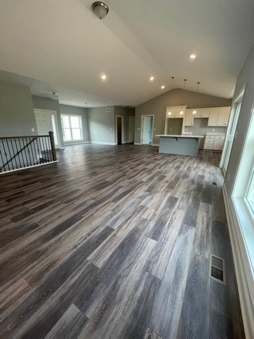 Open-concept room with hardwood flooring, modern kitchen featuring light cabinetry and countertops, visible staircase with wood treads, recessed ceiling lights, and wall vent