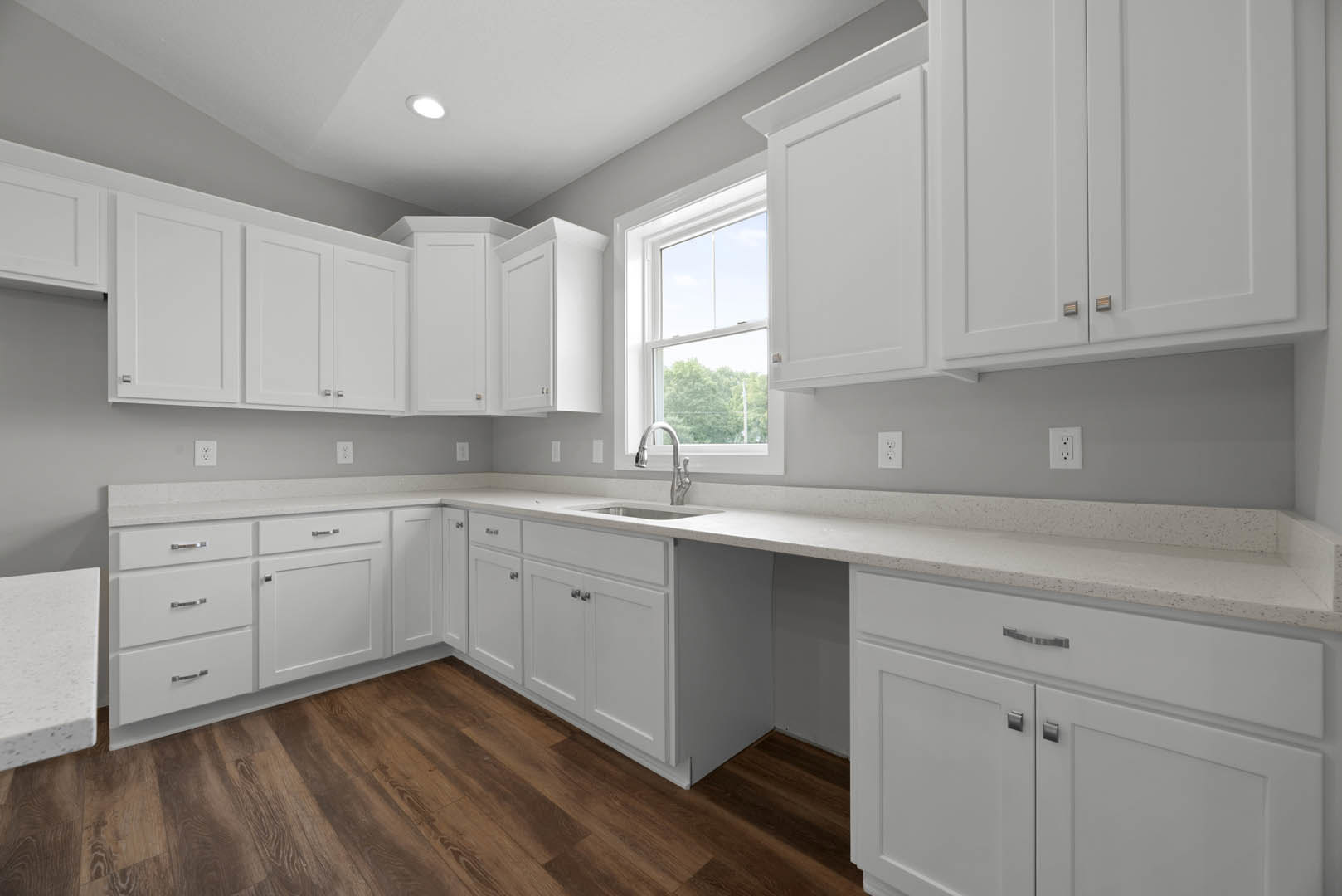 White kitchen cabinets and drawers, speckled white countertop with silver faucet, wood flooring, window overlooking trees.