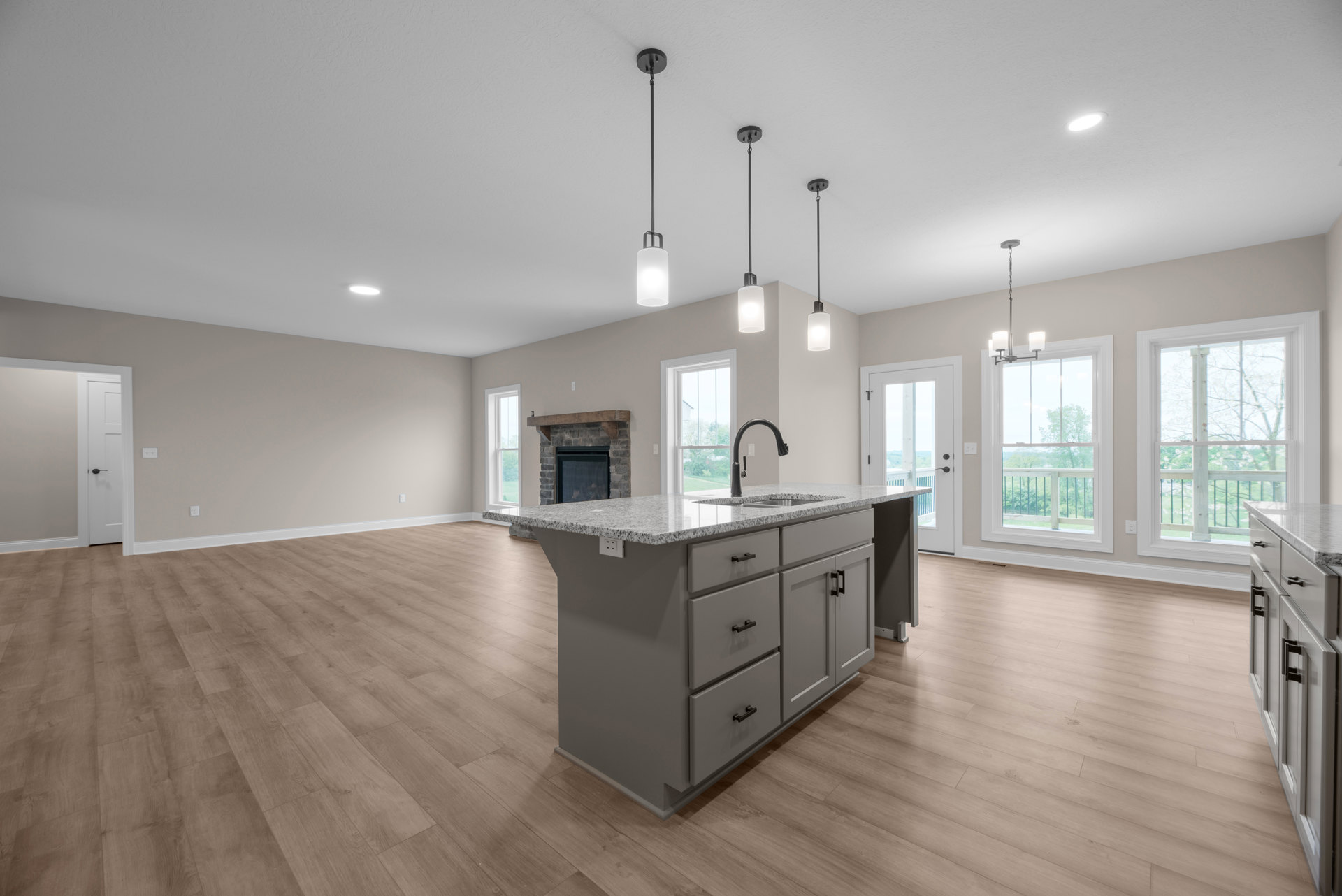 Open-concept kitchen featuring a central island with sink, light wood cabinetry, built-in fireplace, large windows overlooking trees, and hardwood flooring