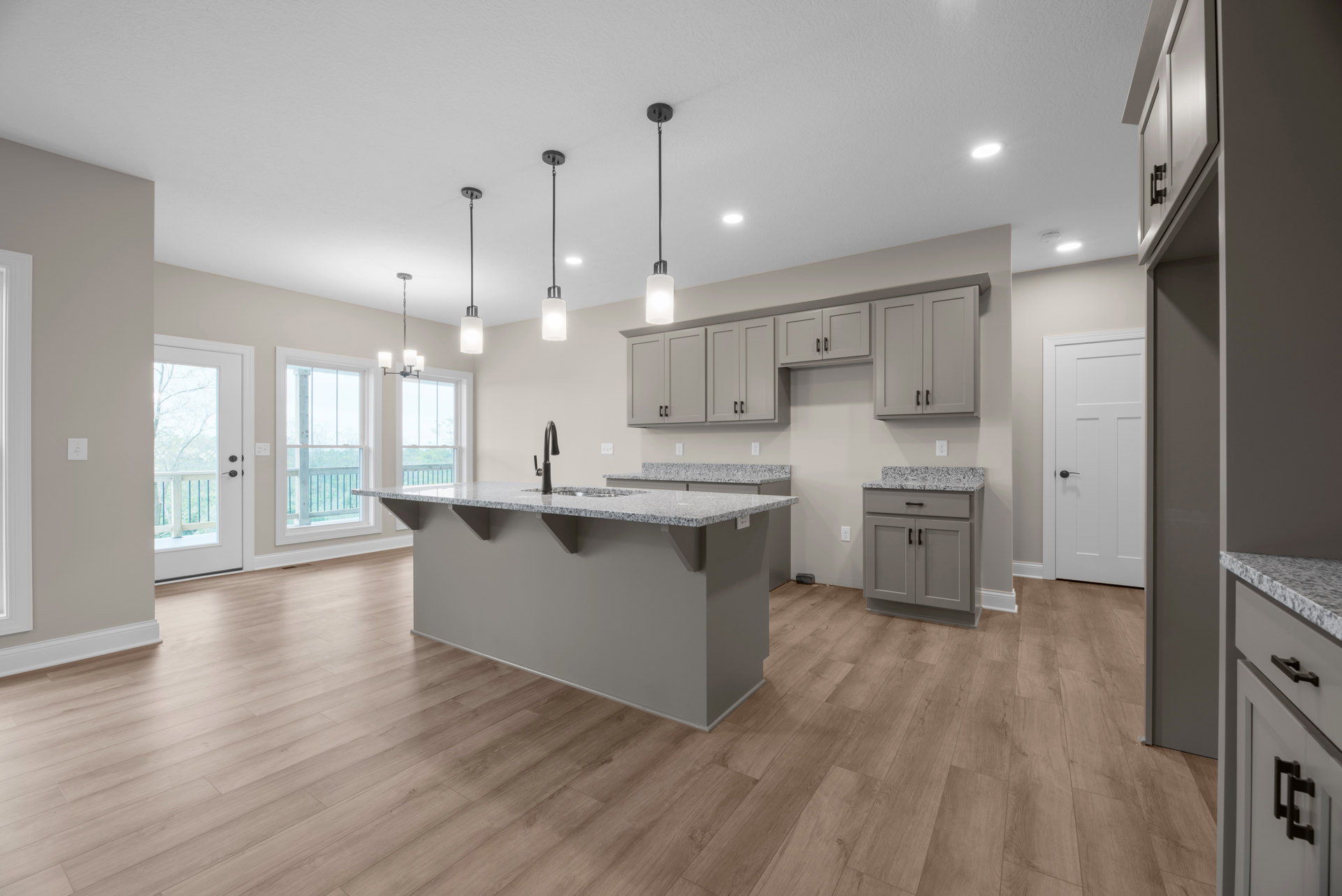 Kitchen with light wood flooring, white cabinetry, stone countertop with stainless steel sink, white door with black handle, large window overlooking outdoor greenery