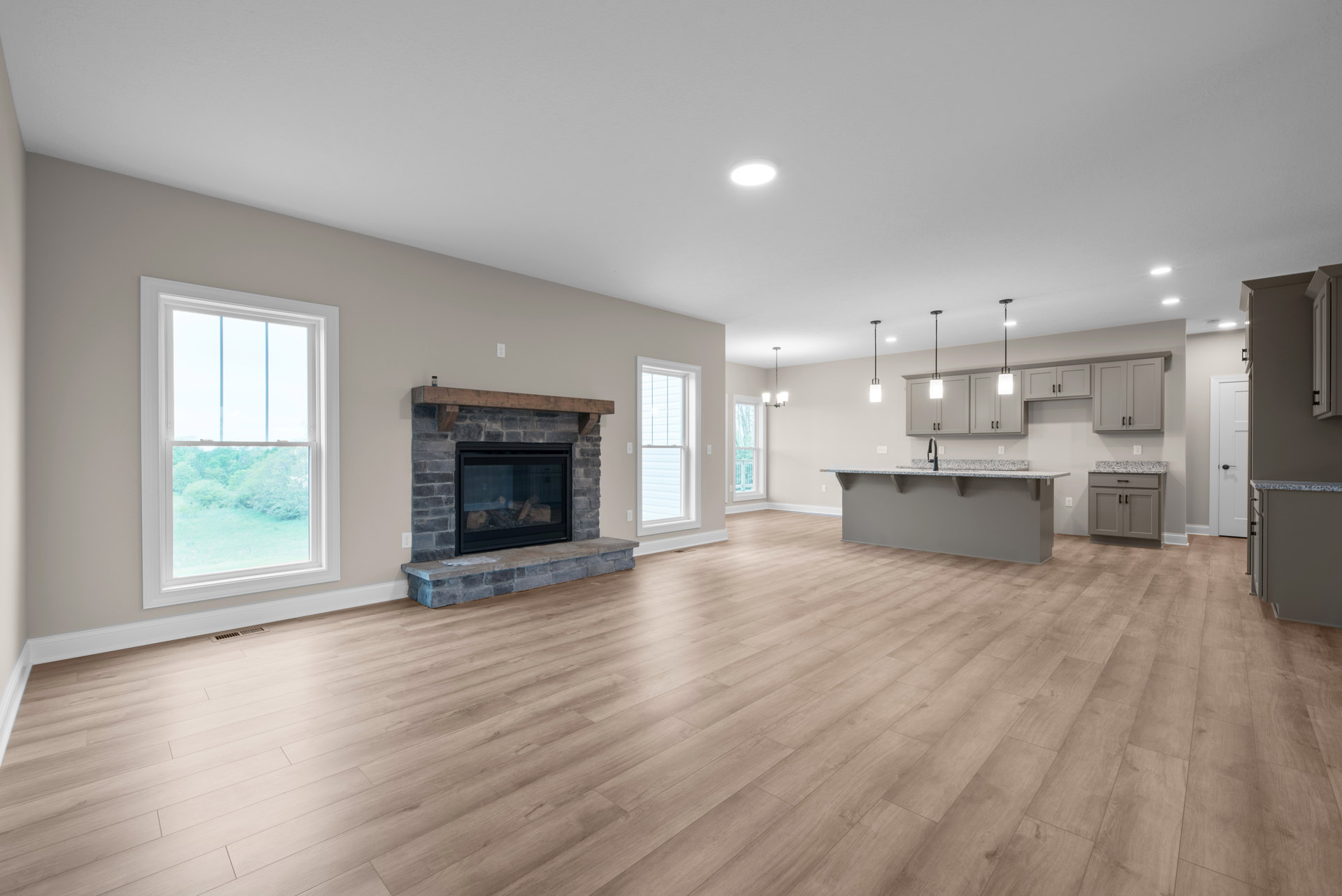 Open concept living room with hardwood floors, stone fireplace with glass door, recessed ceiling light, adjacent kitchen with wood cabinetry, large window showing trees outside