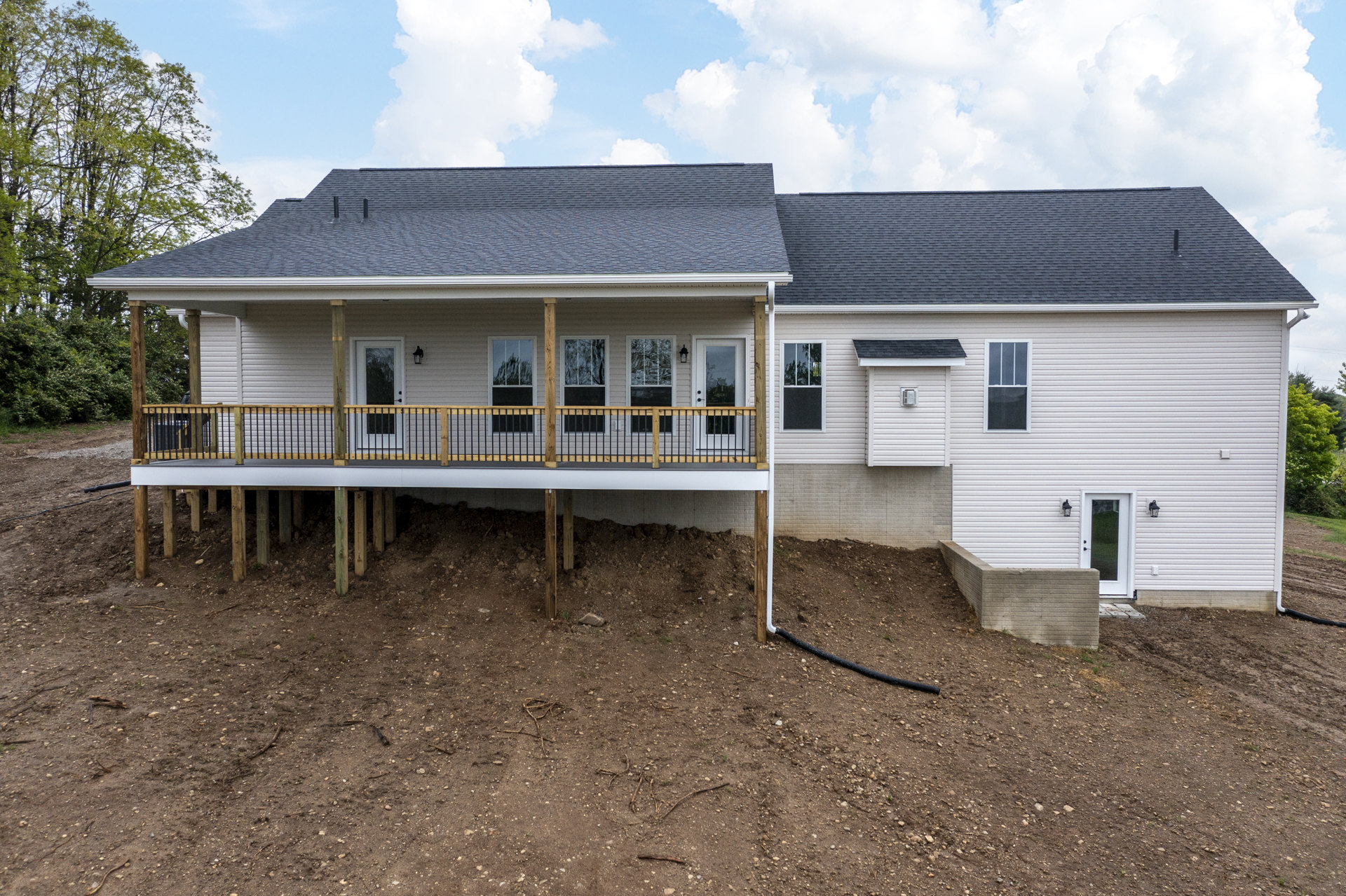 Two-story cottage with wood deck and covered front porch, white siding, screened window, and concrete foundation, surrounded by trees and lawn.