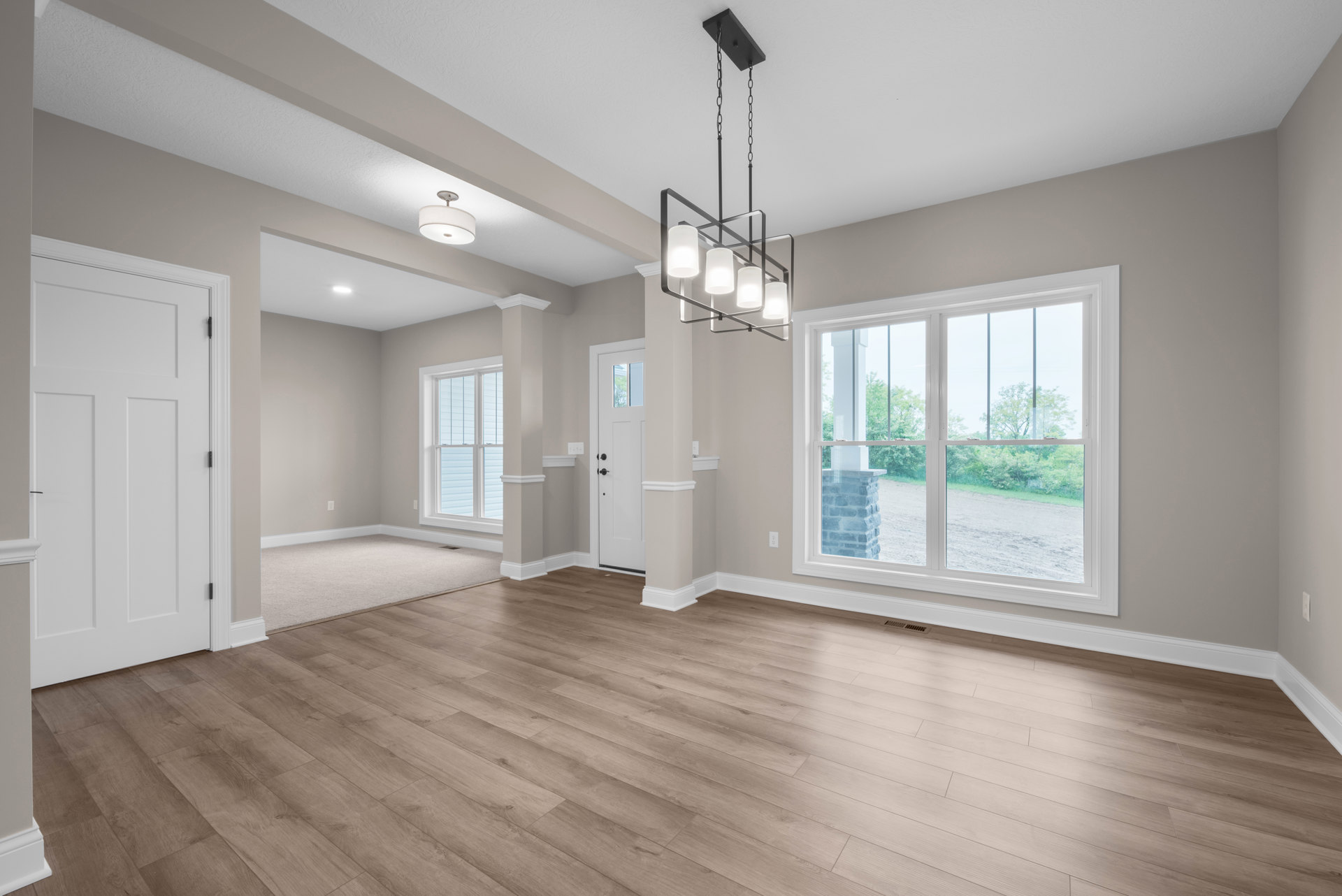 Wood flooring with white baseboards, white door featuring a black handle, window framed by a brick column and stone pillar, ceiling-mounted chandelier with white lamps.