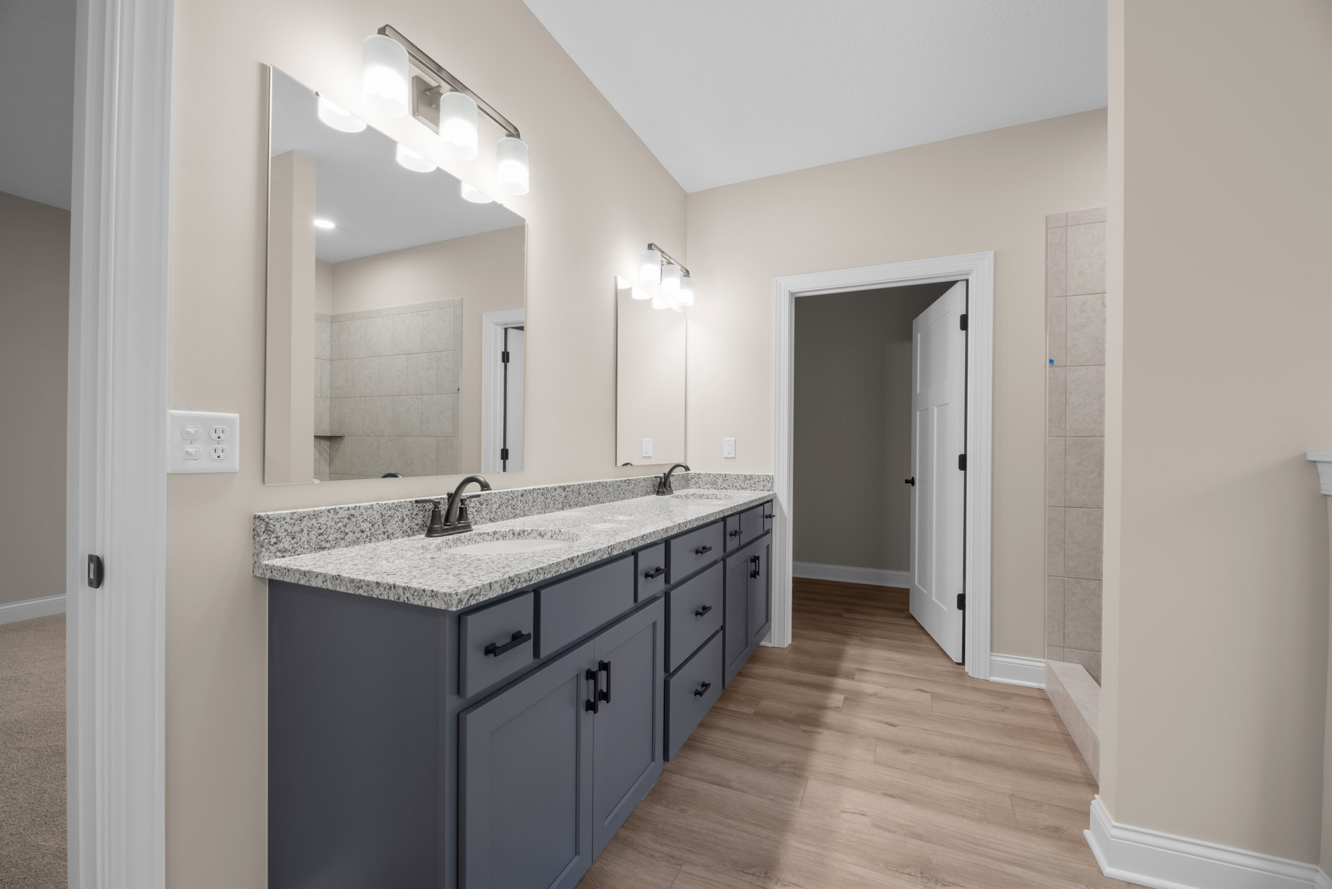 Bathroom with double vanity featuring white sinks, wide mirror above, wood cabinetry and drawers below, tile flooring, wall outlet, and glass-enclosed shower in background.